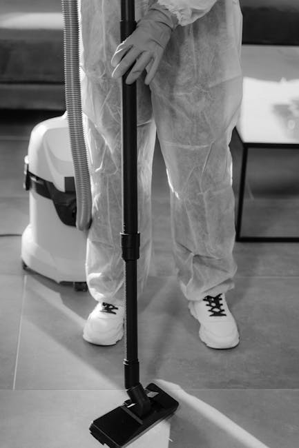 A person dressed in protective coveralls, gloves, and sneakers operating a vacuum cleaner with a long floor nozzle on a smooth, polished surface. The scene appears to be in a residential or commercial